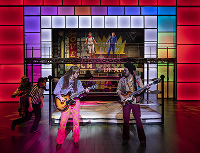 Two guitarists in 1960s clothing play in front of a wrestling ring, and colorful lit panels framing a montage of neon signs. Referee running around to dismantle the ring.